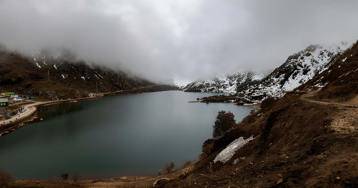 A dramatic, moody view of the sacred Tsomgo (Changu) Lake in Sikkim, featuring dark glacial waters surrounded by rugged, snow-dusted mountains partially hidden by heavy, low-hanging clouds.
