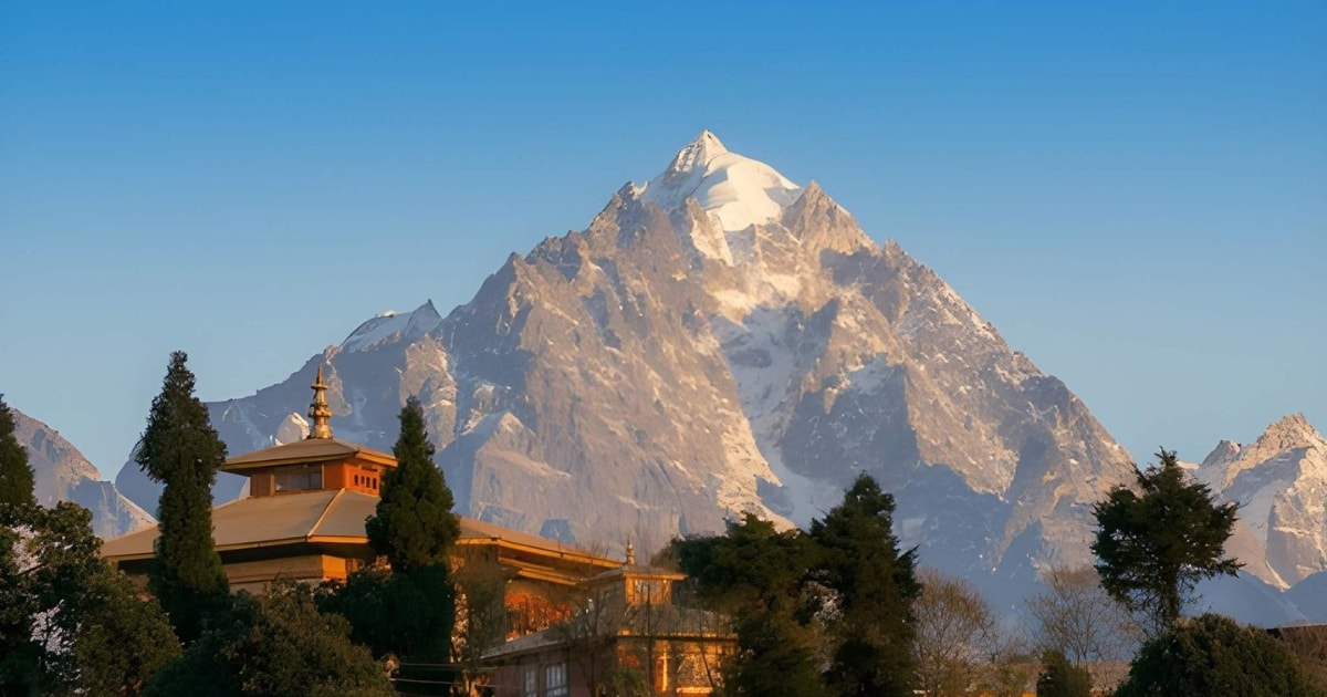 A traditional building with a tiered golden roof sits nestled among dark green trees in the foreground, contrasting sharply with the massive, snow-covered peak of Mount Siniolchu, which is glowing warmly in the sunrise light against a clear blue sky.