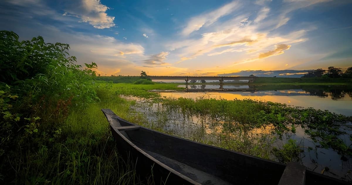 A rustic wooden boat moored in the lush green wetlands of Majuli Island, Assam, overlooking a calm river that perfectly reflects a vibrant, cloudy sunset and a distant bridge.