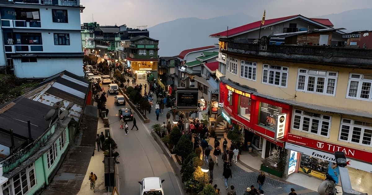 An elevated evening view of MG Marg in Gangtok, a bustling, vehicle-free pedestrian street lined with colorful shops, restaurants, and strolling tourists, set against a mountainous twilight backdrop.