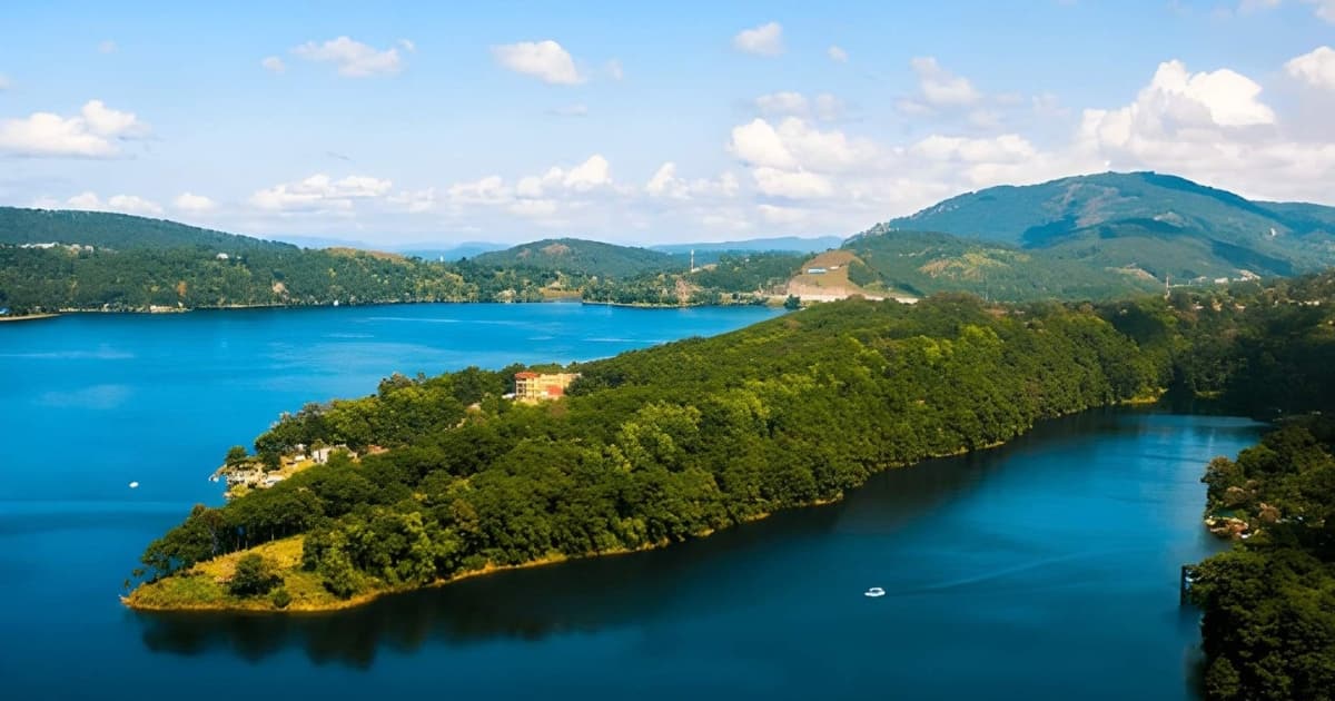 An elevated, panoramic view of a bright blue lake. A large, thickly forested peninsula stretches out into the water, with rolling green mountains under a blue sky in the background.